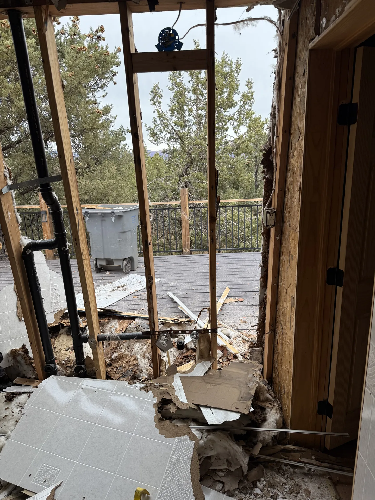 Structural bathroom remodel — interior view looking out through removed exterior wall, framing exposed, plumbing rough-in visible