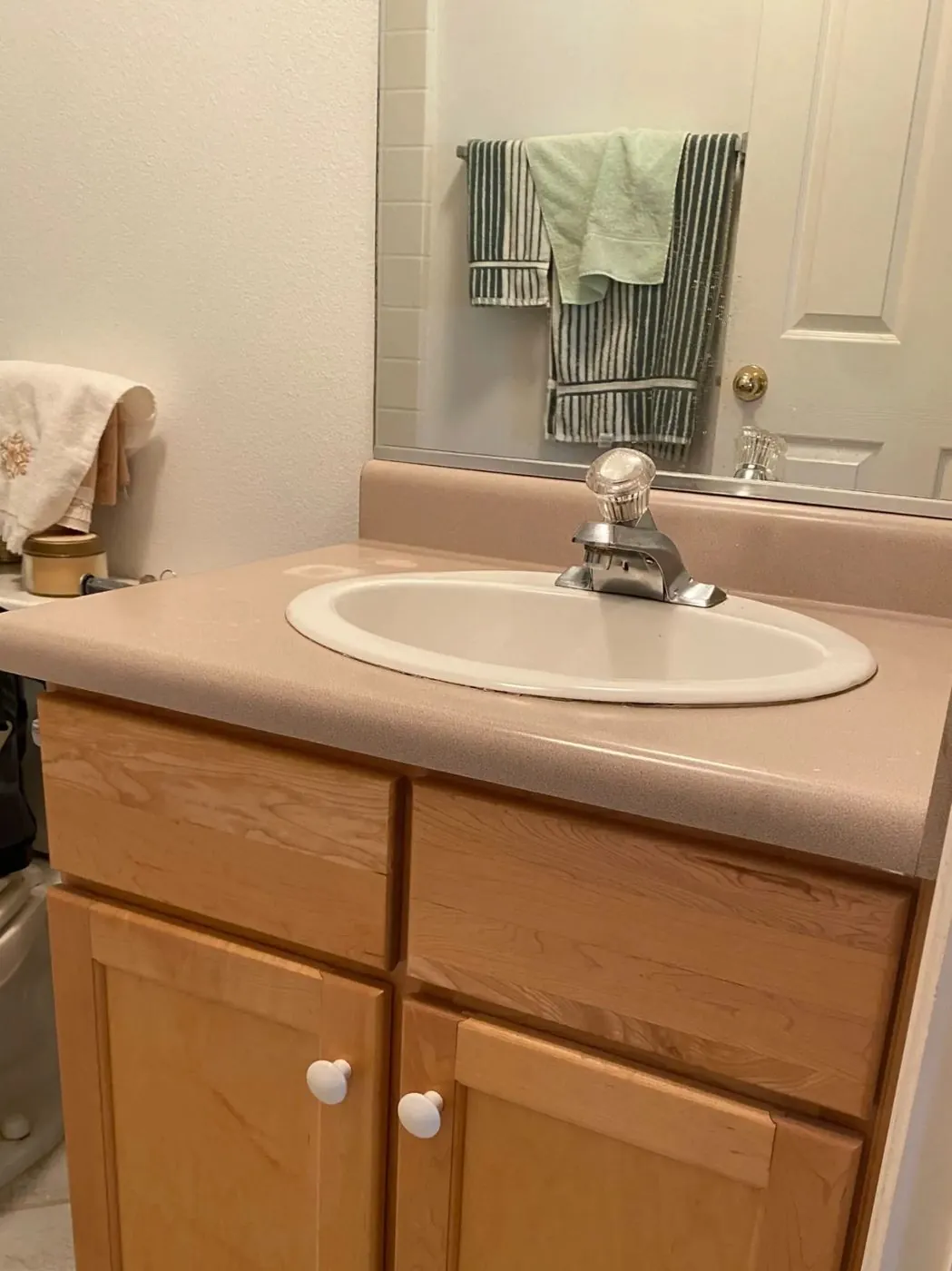 Bathroom vanity before faucet replacement — old pink countertop with dated crystal-knob faucet, light oak cabinet