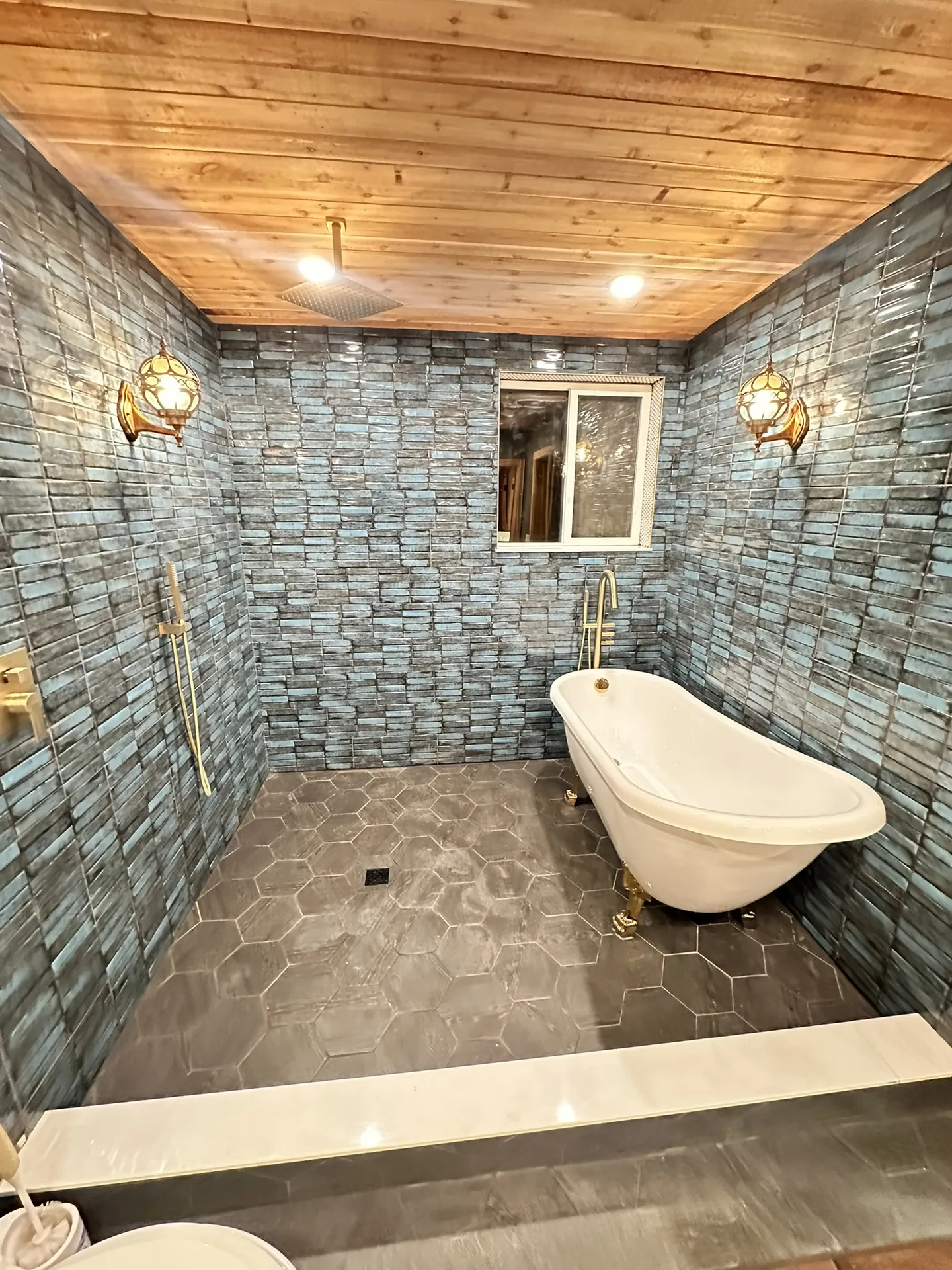 Finished bathroom — rain shower head in cedar ceiling, freestanding tub centered, both sconces lit, full tile walls