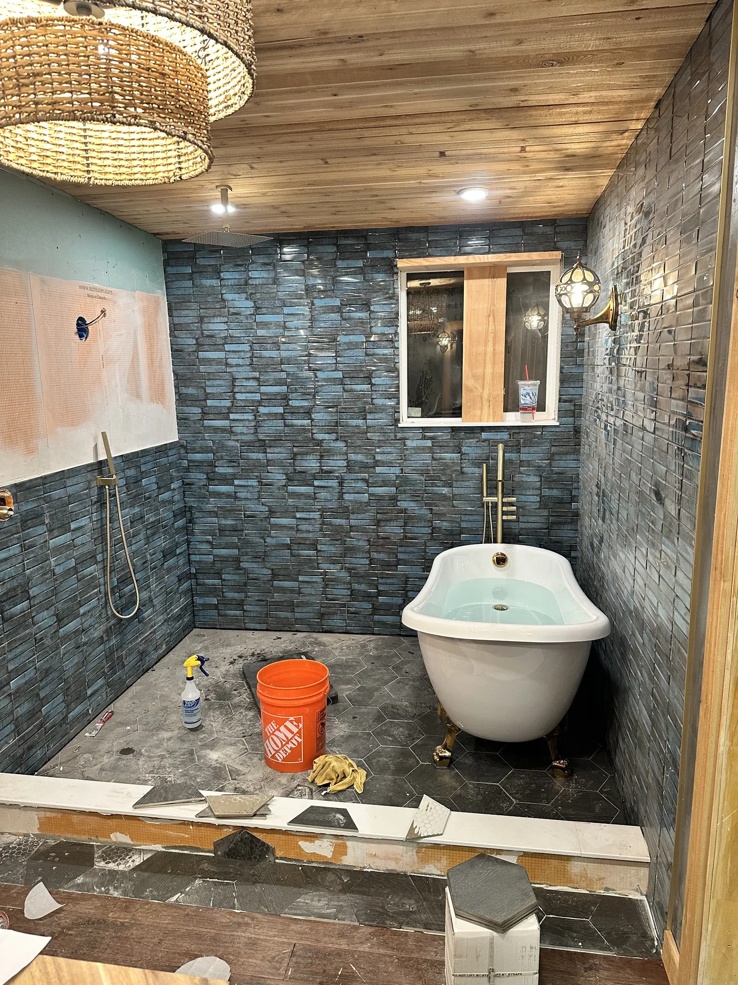 Bathroom remodel progress — near-complete tub room with full blue teal stacked tile walls, clawfoot tub, hexagon floor tile, and chandelier installed