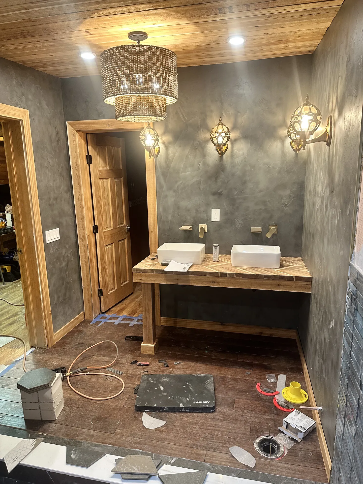 Bathroom remodel progress — vanity area with wood herringbone top, double vessel sinks placed, Moroccan brass sconces lit, Venetian plaster walls, cedar ceiling, worker with tablet visible