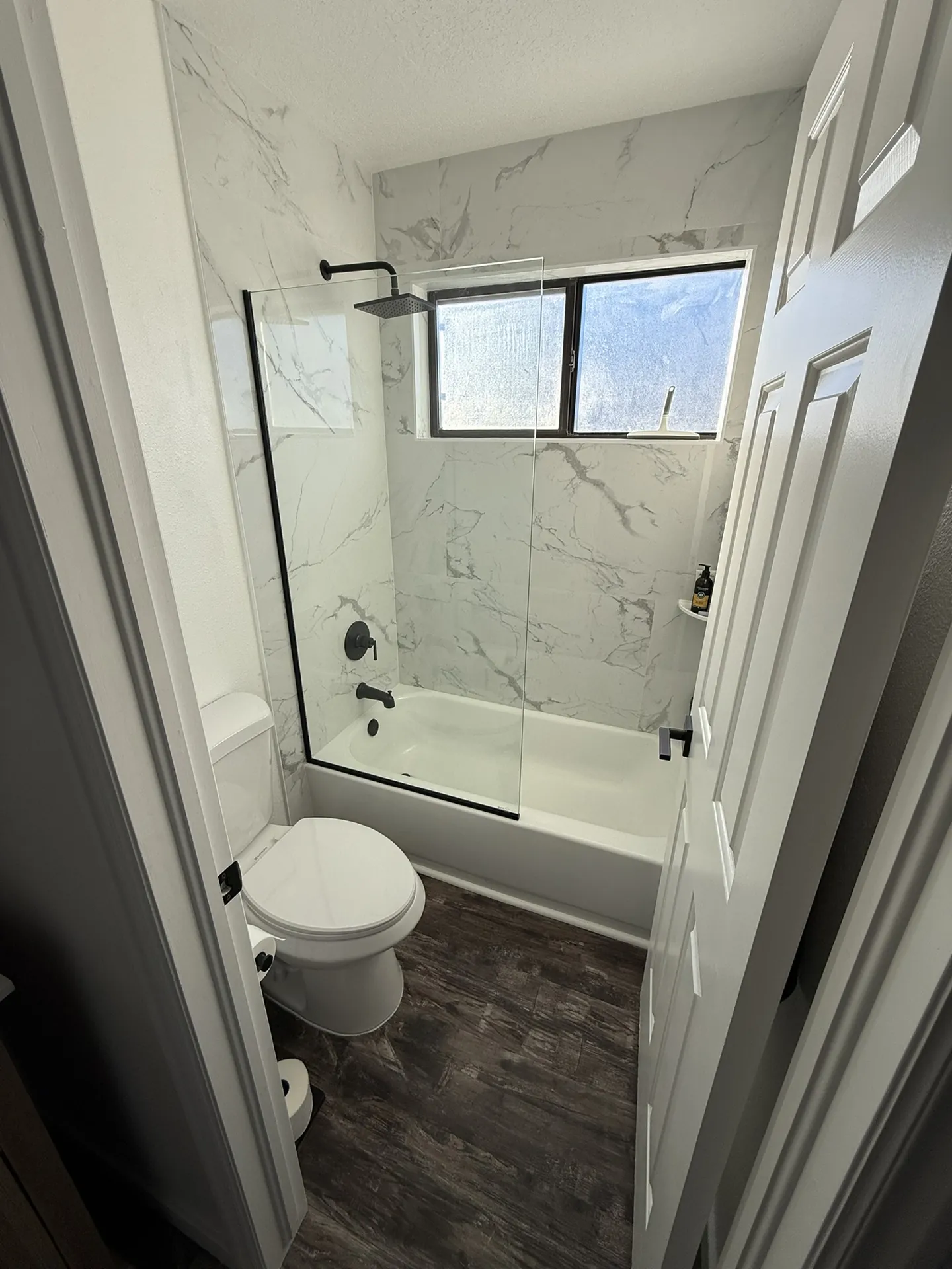 Finished condo — hall bath shower with large-format marble-look tile, glass door, black rain showerhead, and dark LVP floor