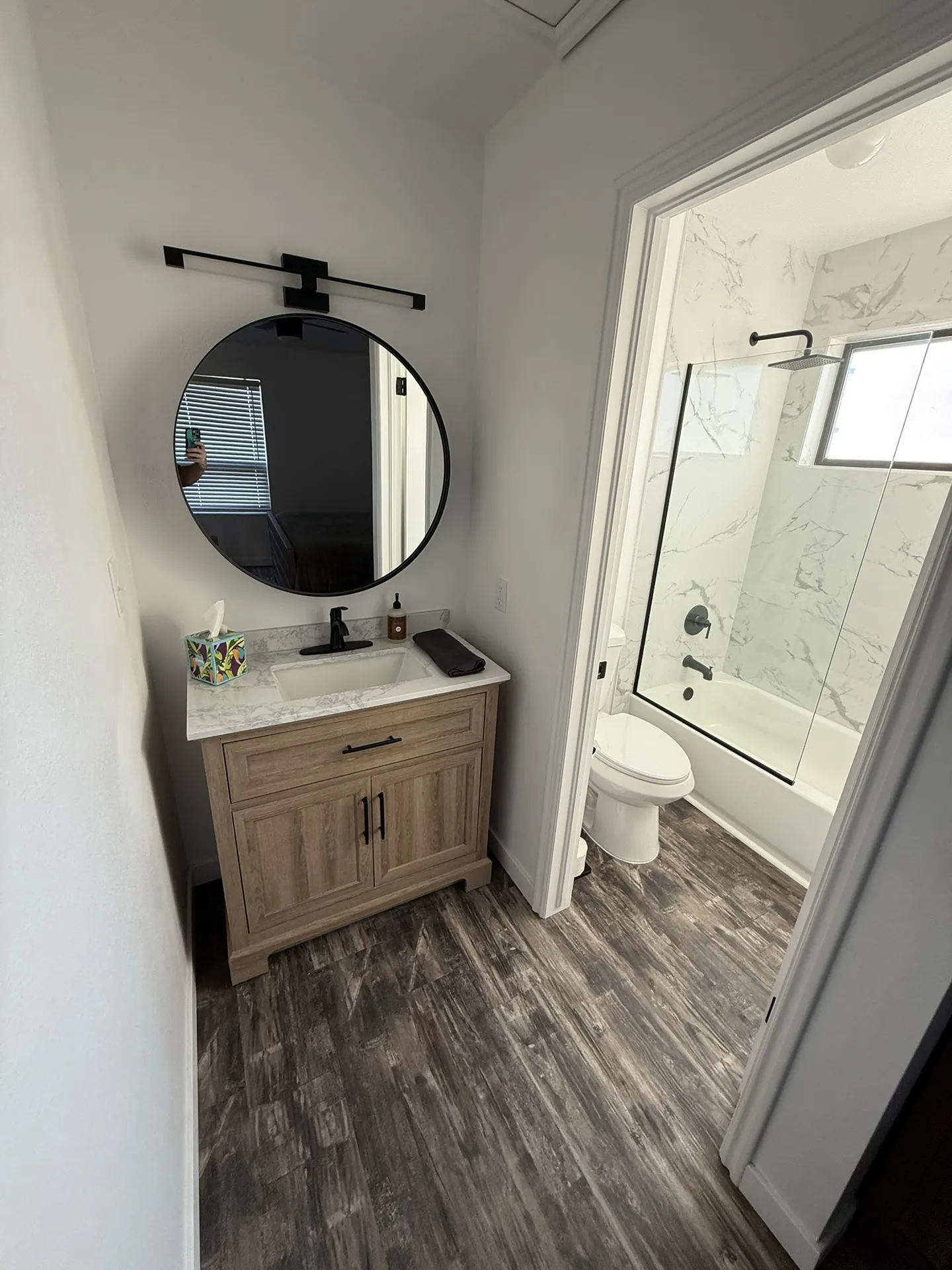 Condo after remodel — hall bathroom with new wood vanity, marble quartz top, round black mirror, black bar light, and new tile shower visible through doorway