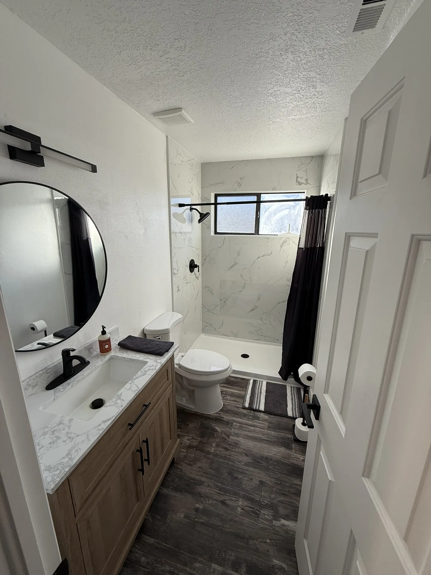 Condo after remodel — master bathroom with new wood vanity, marble quartz, round mirror, walk-in shower with marble tile, and dark LVP floors